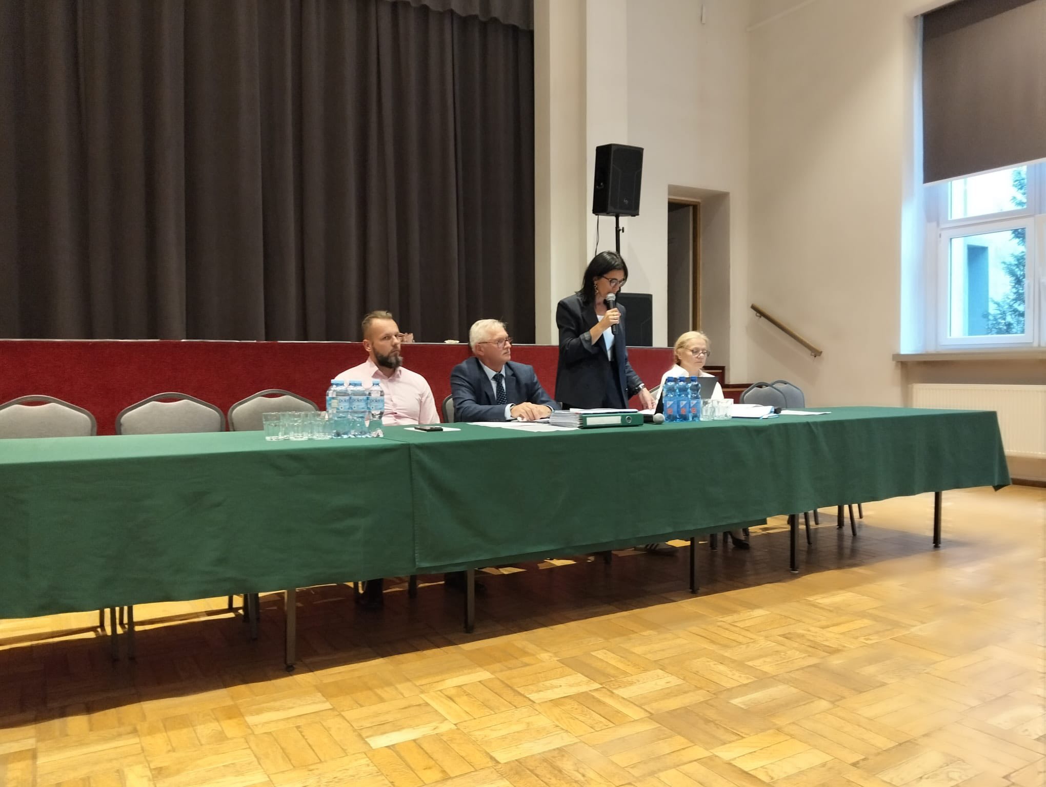 Zdjęcie przedstawia W&oacute;jta gminy, panią Małgorzatę Bednarek zabierającą głos na&nbsp;zebraniu wiejskim. Przy stole siedzą zaproszeni goście i&nbsp;sołtys.